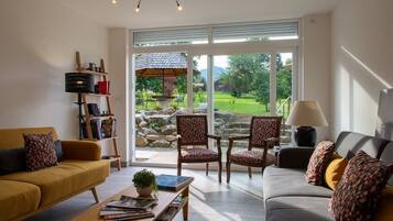 Ein Wohnzimmer mit zwei Sofas, einem Couchtisch und einem Bücherregal. Ein großes Fenster bietet Blick auf einen Garten und eine Terrasse.