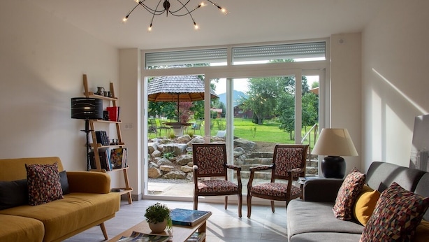 Un salon avec deux canapés, une table basse et une bibliothèque. Une grande fenêtre offre une vue sur un jardin et une terrasse.