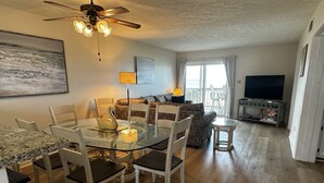A dining area with a glass table, chairs, and a swan-shaped centerpiece. There is a ceiling fan, a sofa, and a television in the background. - Oceanfront 2BR/2 Bath Second Floor Condo with Pool and Oceanfront Deck (Rodanthe)