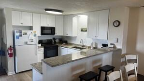 A kitchen with white cabinets, a black microwave, a stove, a sink, and a small island with bar stools. - Oceanfront 2BR/2 Bath Second Floor Condo with Pool and Oceanfront Deck (Rodanthe)