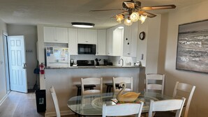 A modern kitchen with white cabinets, a microwave, and a dining area with a glass table and white chairs. - Oceanfront 2BR/2 Bath Second Floor Condo with Pool and Oceanfront Deck (Rodanthe)