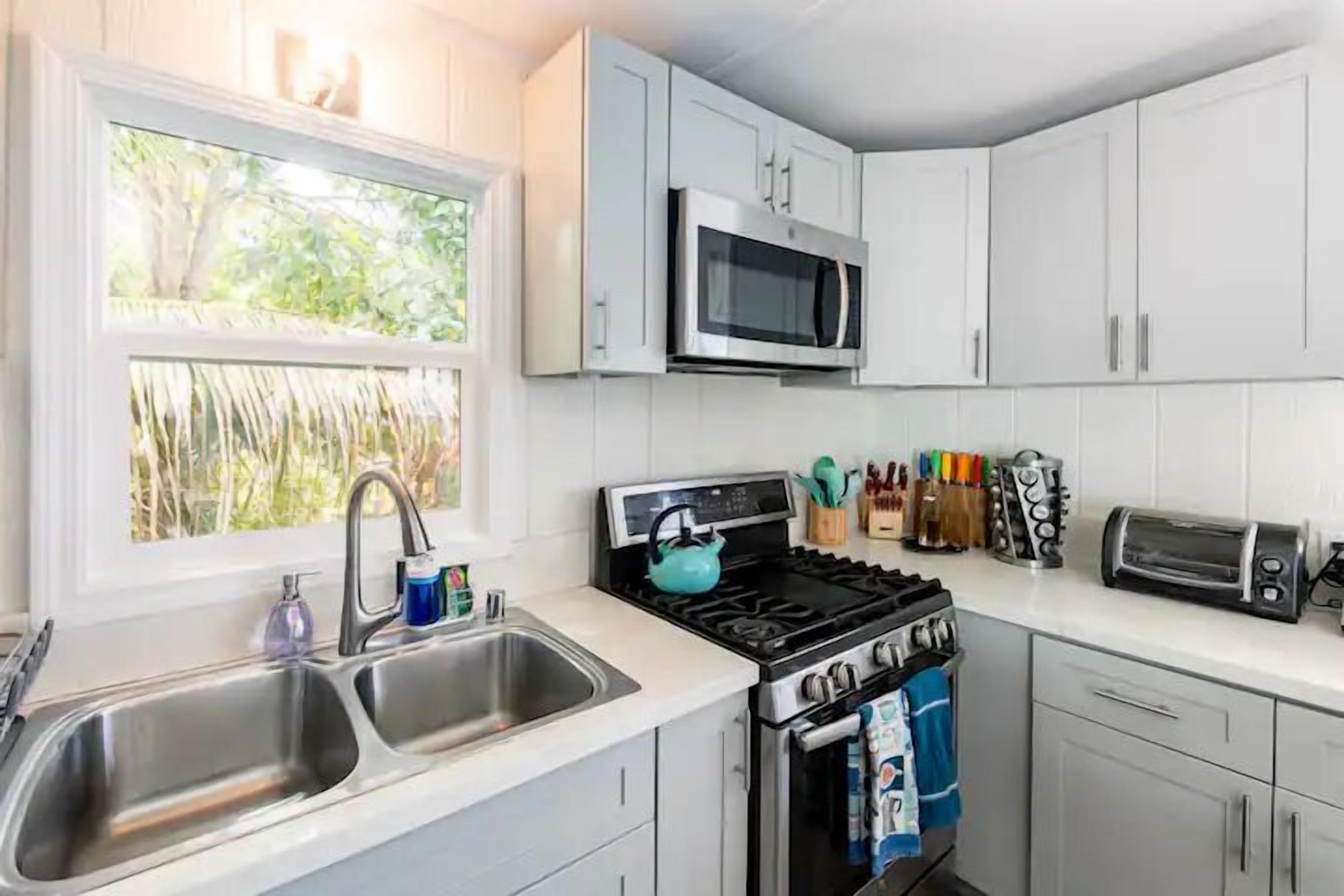 A modern kitchen with white cabinets, a stainless steel sink, a stove, a microwave, and a window with a view of greenery.