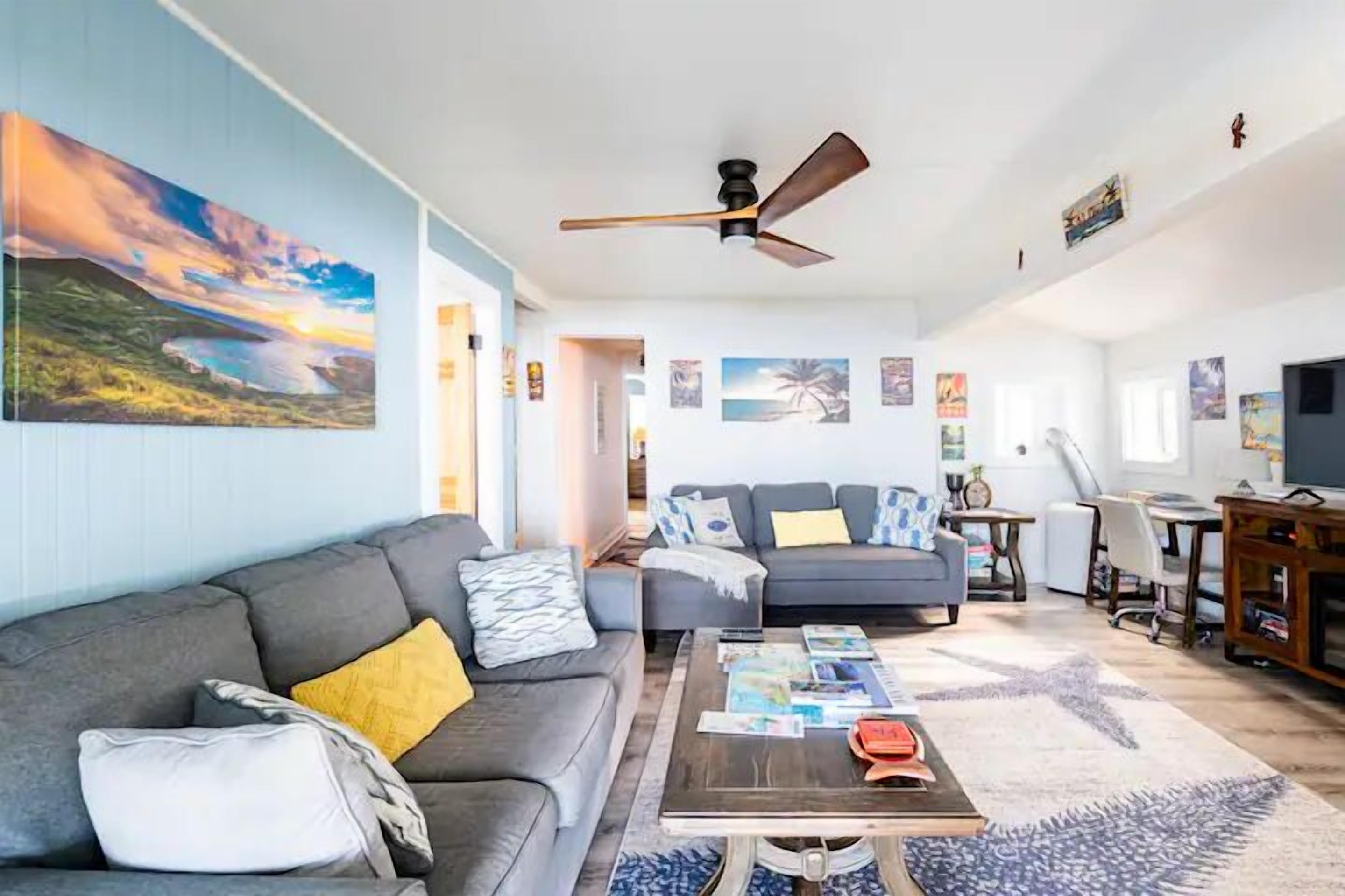 A spacious living area with a gray sofa, a coffee table, and a ceiling fan. There are multiple paintings on the walls and a TV on a wooden stand.