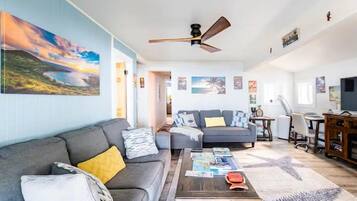 A spacious living area with a gray sofa, a coffee table, and a ceiling fan. There are multiple paintings on the walls and a TV on a wooden stand.
