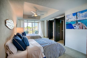 A hotel room with two beds, a ceiling fan, a window with curtains, a painting of a sailboat, and a door leading to another room.