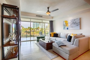 A modern living room with a grey sofa, a ceiling fan, and a large window with a view of palm trees and buildings.