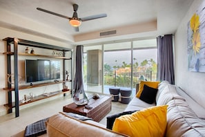 A modern living room with a flat-screen TV, a ceiling fan, a large window with a view of palm trees, and a sofa with yellow cushions.
