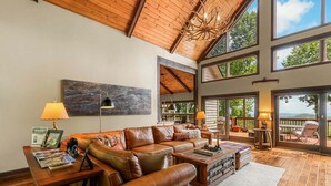 A spacious living room with a leather sofa, a coffee table, and a chandelier. There are large windows and a view of the outdoors. - Discover Bear Trail in Boone, NC—a cozy 4-bedroom chalet perfect for 8 guests. (Boone)