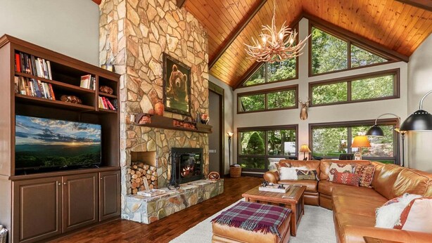 A spacious living room with a stone fireplace, a television, leather furniture, and large windows. - Discover Bear Trail in Boone, NC—a cozy 4-bedroom chalet perfect for 8 guests. (Boone)