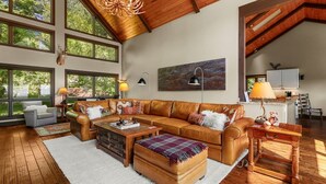 A spacious living room with a large brown leather sofa, a wooden coffee table, and a fireplace. There are large windows allowing natural light and views of the outdoors. - Discover Bear Trail in Boone, NC—a cozy 4-bedroom chalet perfect for 8 guests. (Boone)