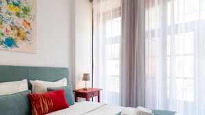 A neatly made bed with a teal bedspread, white pillows, and a red accent pillow. There is a small bedside table with a lamp and a folded towel on the bed.