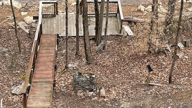 Eine Holzsteg, der zu einer kleinen Holzplattform mit Metallgeländer führt, umgeben von Bäumen und einem Gewässer.