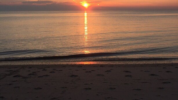 Ein Strand mit Fußspuren, die zum Wasser führen, ein Sonnenuntergang im Hintergrund und ruhige See.