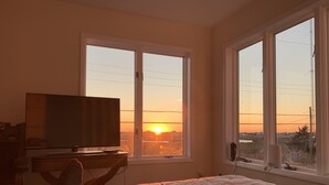 Ein Schlafzimmer mit Bett, Fernseher auf einem Schrank und großen Fenstern mit Blick auf den Sonnenuntergang.