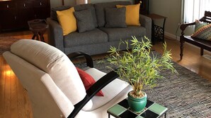 Ein Wohnzimmer mit einer Couch, einem Sessel und einem Couchtisch mit einer Pflanze. Eine Deckenventilator, eine Uhr und ein gerahmtes Bild sind an der Wand zu sehen.