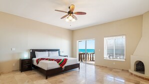A bedroom with a bed, a nightstand, a ceiling fan, a fireplace, and a balcony with a view of the ocean. - The Suites at Rocky Point 2 (Puerto Peñasco)