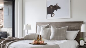 Une chambre moderne avec un grand lit, un tableau représentant un élan accroché au mur, et un plateau avec des bouteilles et des verres posé sur le lit.