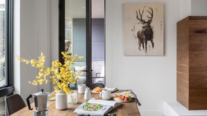 Un coin repas avec une table en bois, une machine à café, un vase avec des fleurs jaunes, une assiette de nourriture et un tableau représentant un cerf accroché au mur.
