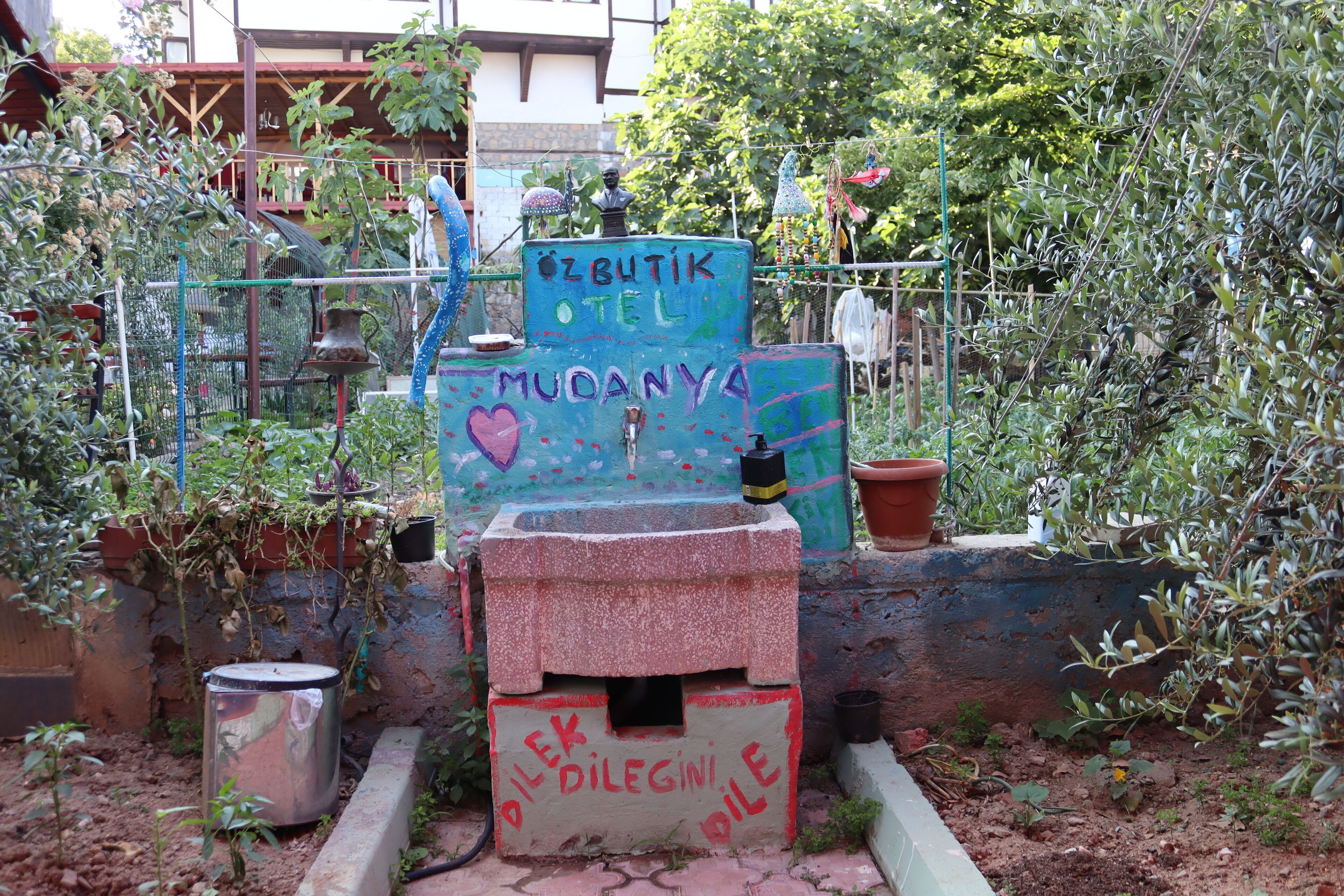 A rustic outdoor area with a fountain, surrounded by plants and a metal fence.