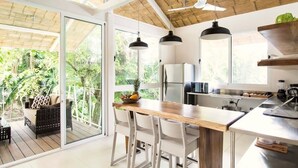 A modern kitchen with a wooden island, white chairs, and a glass door leading to a balcony with a wicker chair and a fruit bowl. - MarAmar Bungalows (Cóbano)