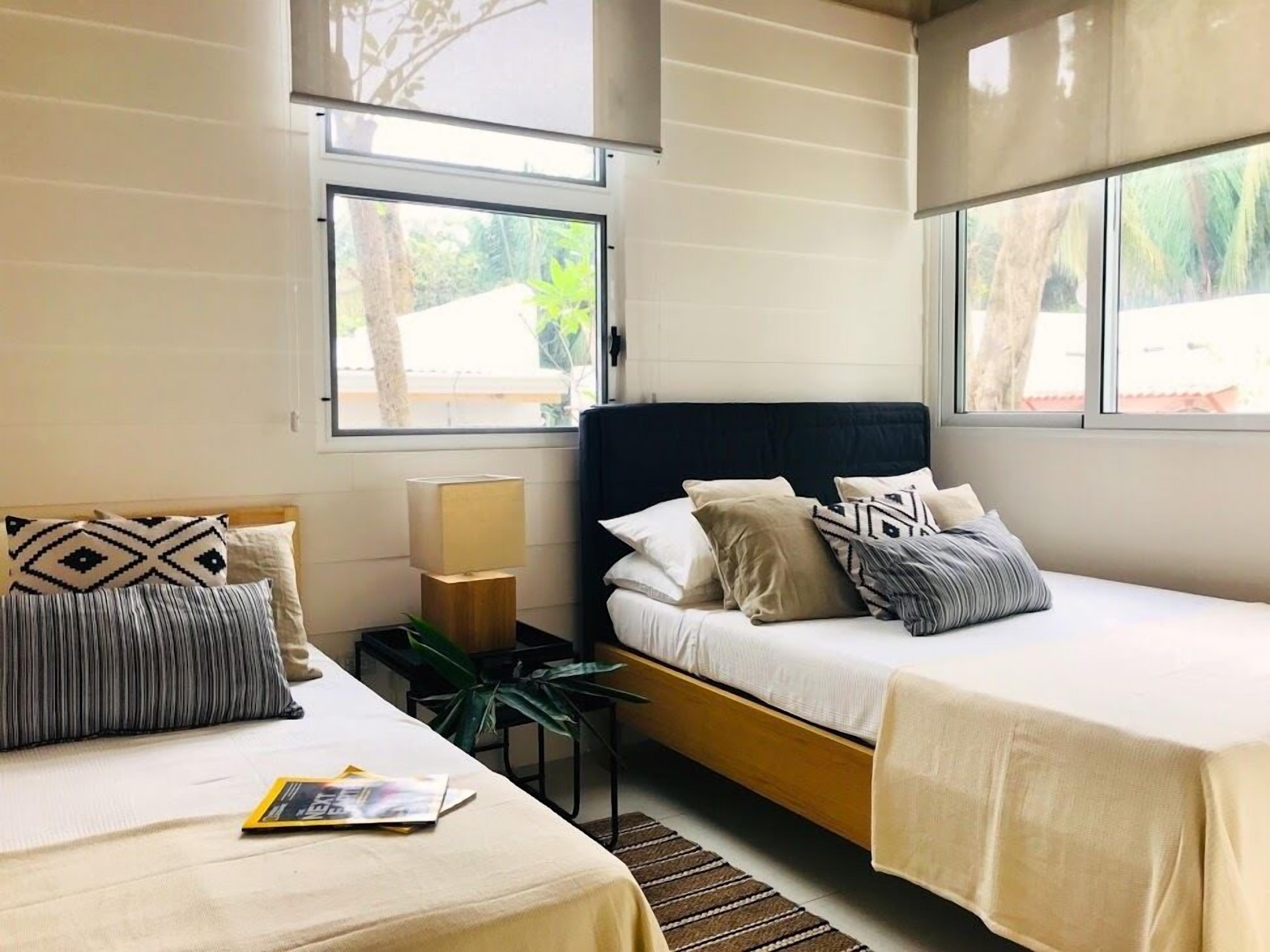 Una habitación de hotel moderna con dos camas, una mesita de noche con lámpara, y ventanas amplias con vistas a zonas verdes.