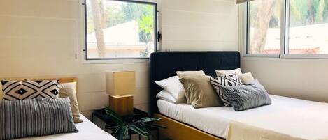 Una habitación de hotel moderna con dos camas, una mesita de noche con lámpara, y ventanas amplias con vistas a zonas verdes.