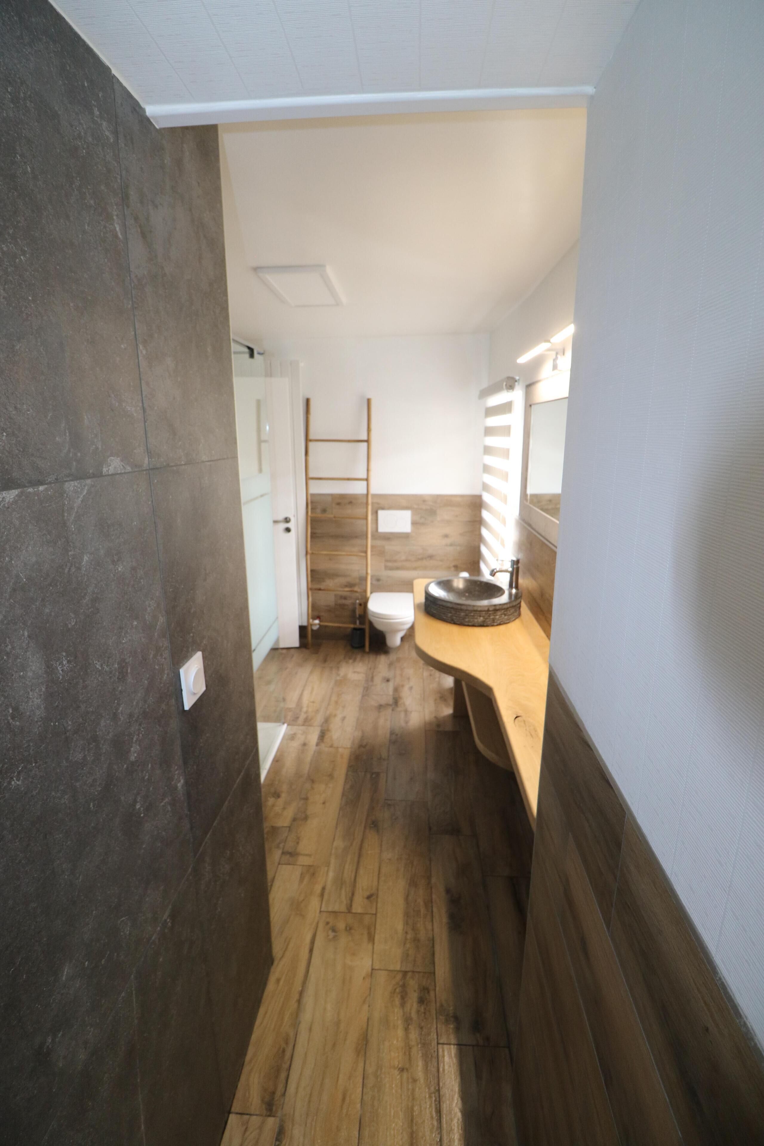 Een smalle gang met een houten vloer die leidt naar een badkamer. De badkamer heeft een houten wastafel met een wastafel en een spiegel.
