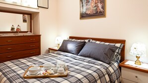 Een net opgemaakt bed met een geruit sprei, een nachtkastje met een lamp, een dressoir met lades en een lijst met een afbeelding aan de muur.