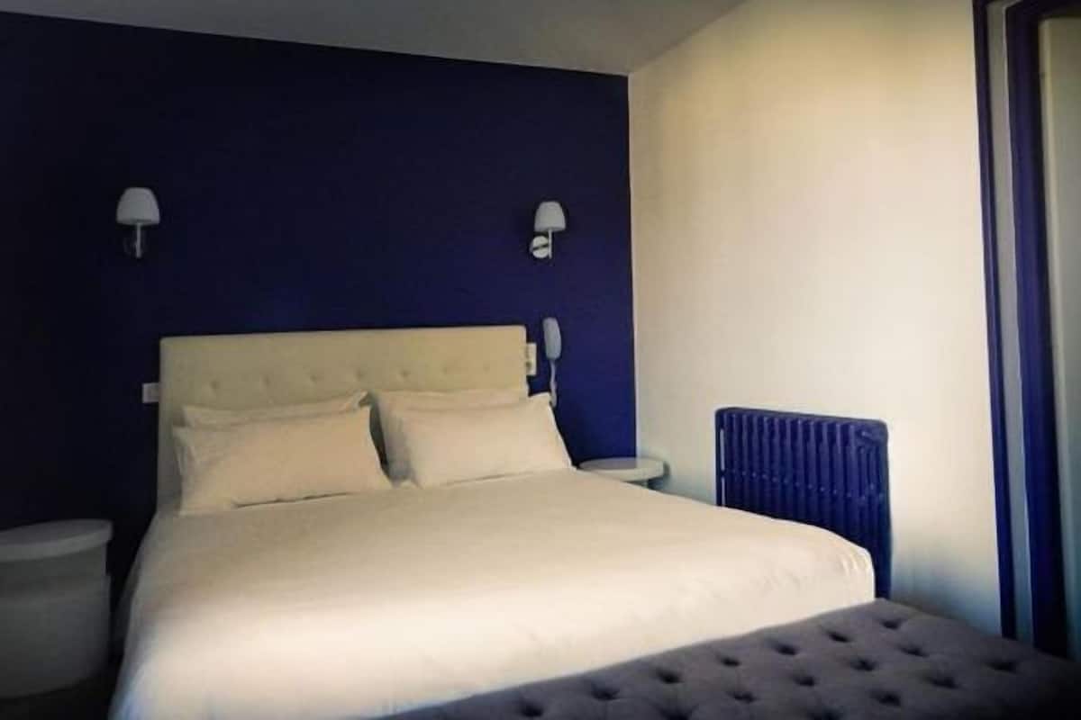 A neatly made bed in a room with a dark blue accent wall, two bedside lamps, and a radiator.