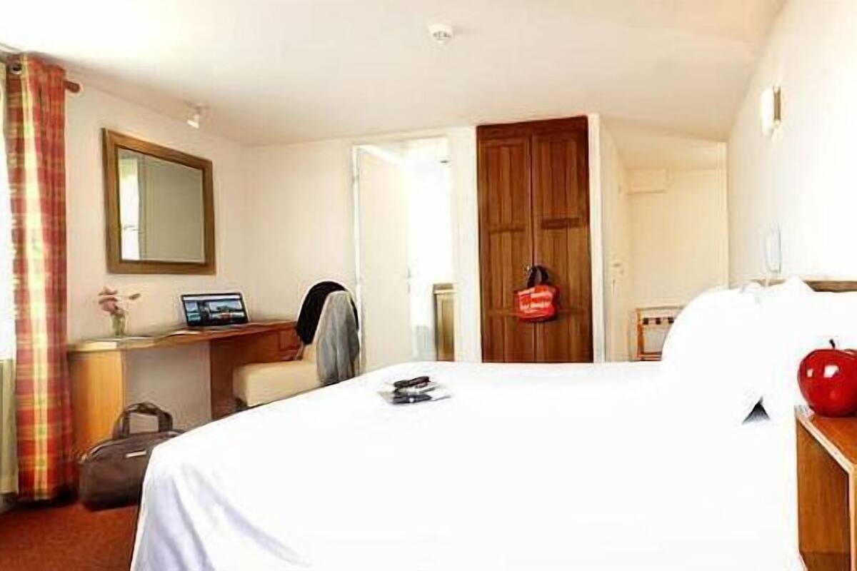 A hotel room with a bed, a desk, a chair, a suitcase, and a red apple on the nightstand.