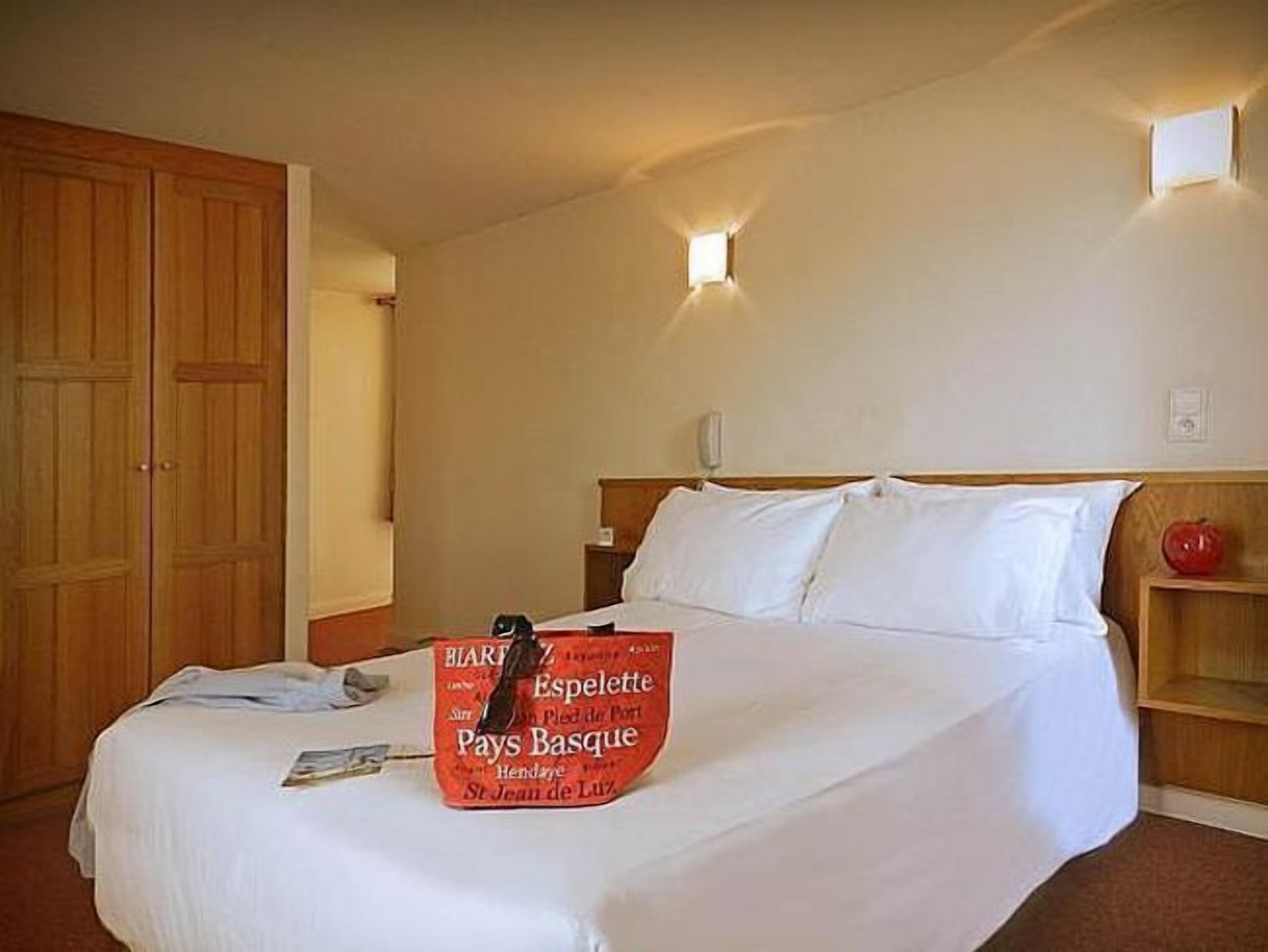 A hotel room with a bed, a wooden wardrobe, a nightstand, and a wall-mounted light fixture.