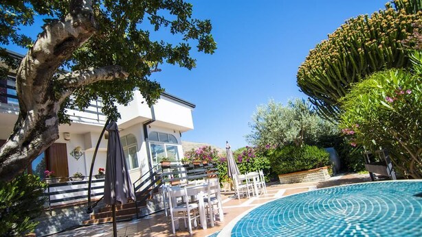 A pool area with a tiled edge, outdoor furniture, and a building in the background. - Grotta dei Puntali (Carini)