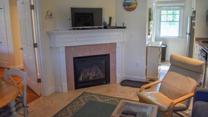 A living room with a fireplace, a television on a mantel, a clock on the wall, and a window with blinds. - Completely updated 1 bedroom condo unit-steps to a shared private beach! (Dennis Port)