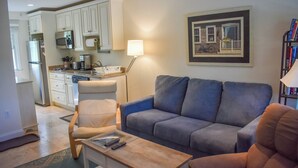 A living room with a grey sofa, a wooden coffee table, and a chair. There is a kitchen in the background with white cabinets and a microwave. - Completely updated 1 bedroom condo unit-steps to a shared private beach! (Dennis Port)
