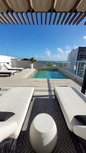 Zona de piscina en la azotea con sillones reclinables y una pequeña mesa redonda, con vistas a un cielo azul despejado y edificios a la distancia.