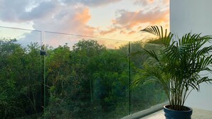 A balcony with a glass railing overlooking a lush green landscape and a sunset sky. - MOB 205 by Riviera Rentals (Playa del Carmen)