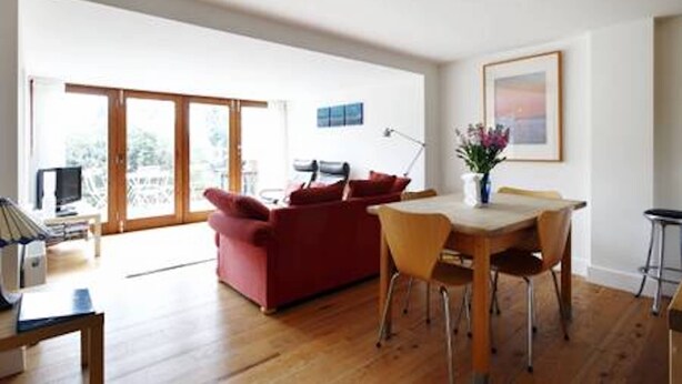 A spacious living area with a red sofa, wooden dining table, and chairs. There is a TV on a stand and a window with a view of greenery.