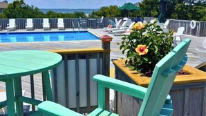 Zona junto a la piscina con una terraza de madera, una mesa redonda y una jardinera con flores.