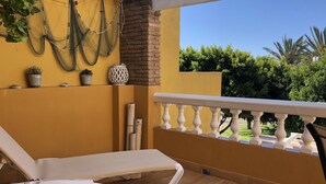 Un balcon avec une chaise blanche, des murs jaunes, un pilier en briques et une vue sur la verdure.