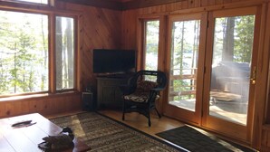 A wooden cabin interior with a television on a cabinet, a chair, and a dining table with a remote control. - Comfortable lake home on pristine Beech Hill Pond (Otis)
