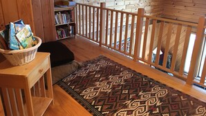 A cozy wooden interior with a bookshelf, a basket of books, a patterned rug, and a wooden railing. - Comfortable lake home on pristine Beech Hill Pond (Otis)