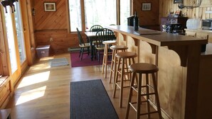 A wooden cabin interior with a kitchen area, dining table, and bar stools. - Comfortable lake home on pristine Beech Hill Pond (Otis)