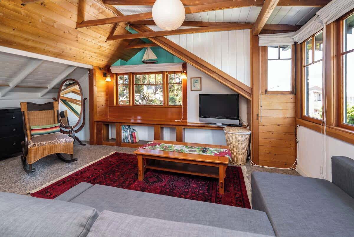 Una habitación cálida en el ático, con vigas de madera, un ventanal, televisor, una cesta de mimbre y una alfombra roja.