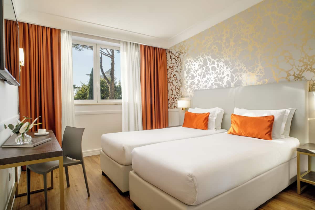 Habitación de hotel con dos camas, un escritorio, una silla, una ventana con cortinas y un papel tapiz decorativo.