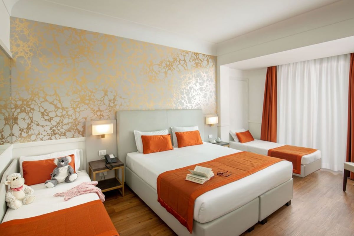 Ein Hotelzimmer mit einem großen Bett, einem kleineren Bett, einem Schreibtisch mit Stuhl, einem Nachttisch und einer Lampe.