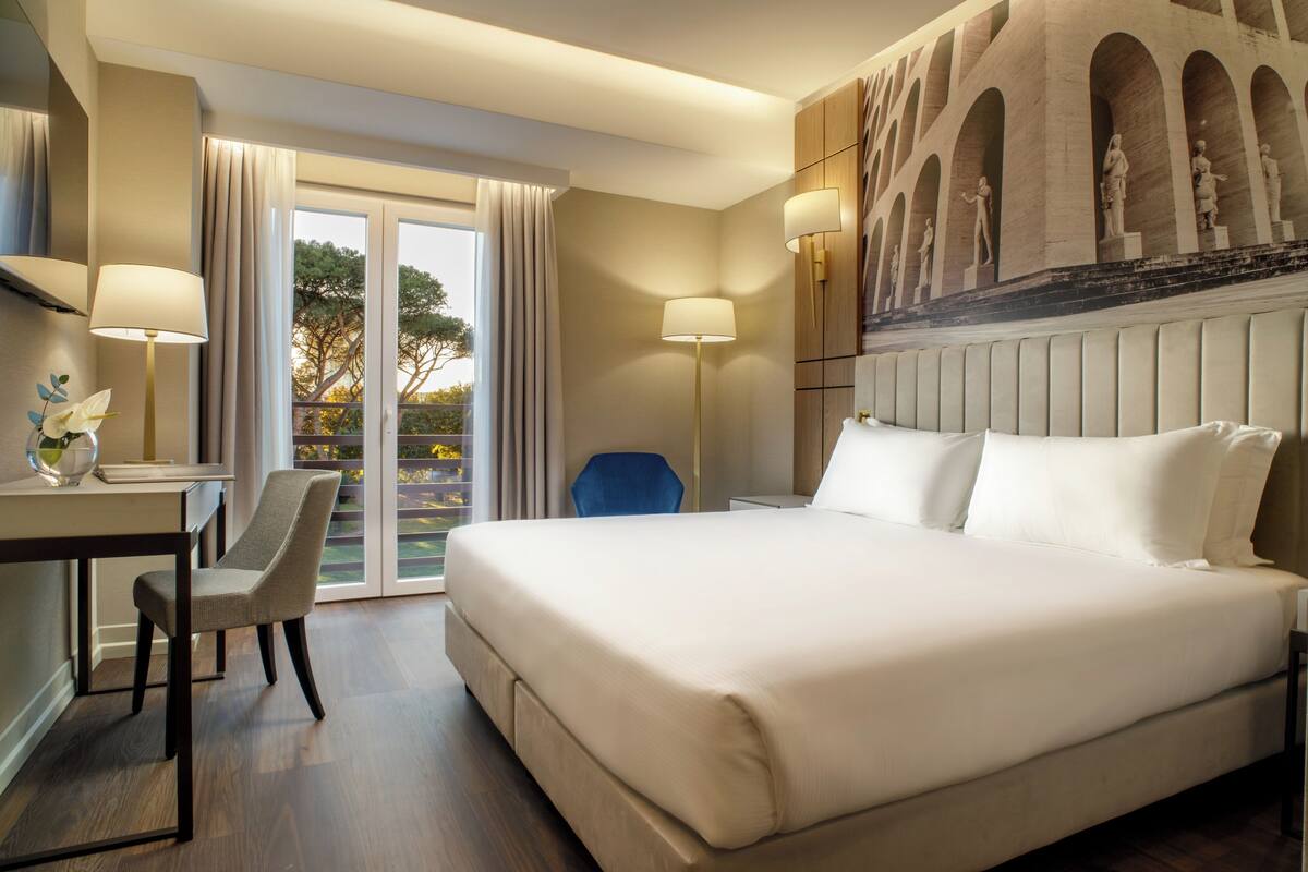 Una habitación de hotel con una cama grande, un escritorio con silla, vistas del exterior a través de un ventanal y una pared con una escultura.
