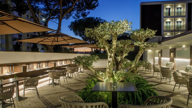 Un área de estar al aire libre con sillas y mesas de mimbre, un árbol en el centro y un edificio con ventanas iluminadas al fondo.