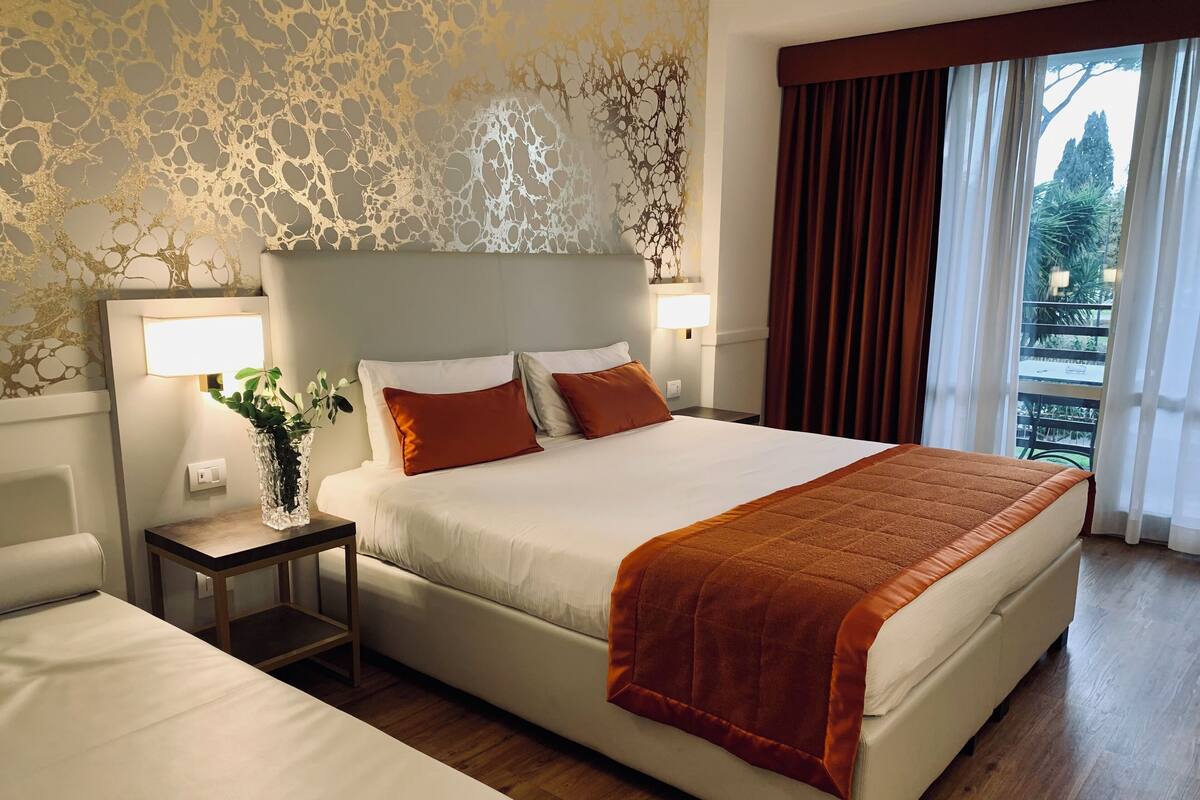 Un cuarto de hotel con una cama grande, una mesita de noche con una lámpara, un florero con una planta y vistas al exterior a través de una ventana.