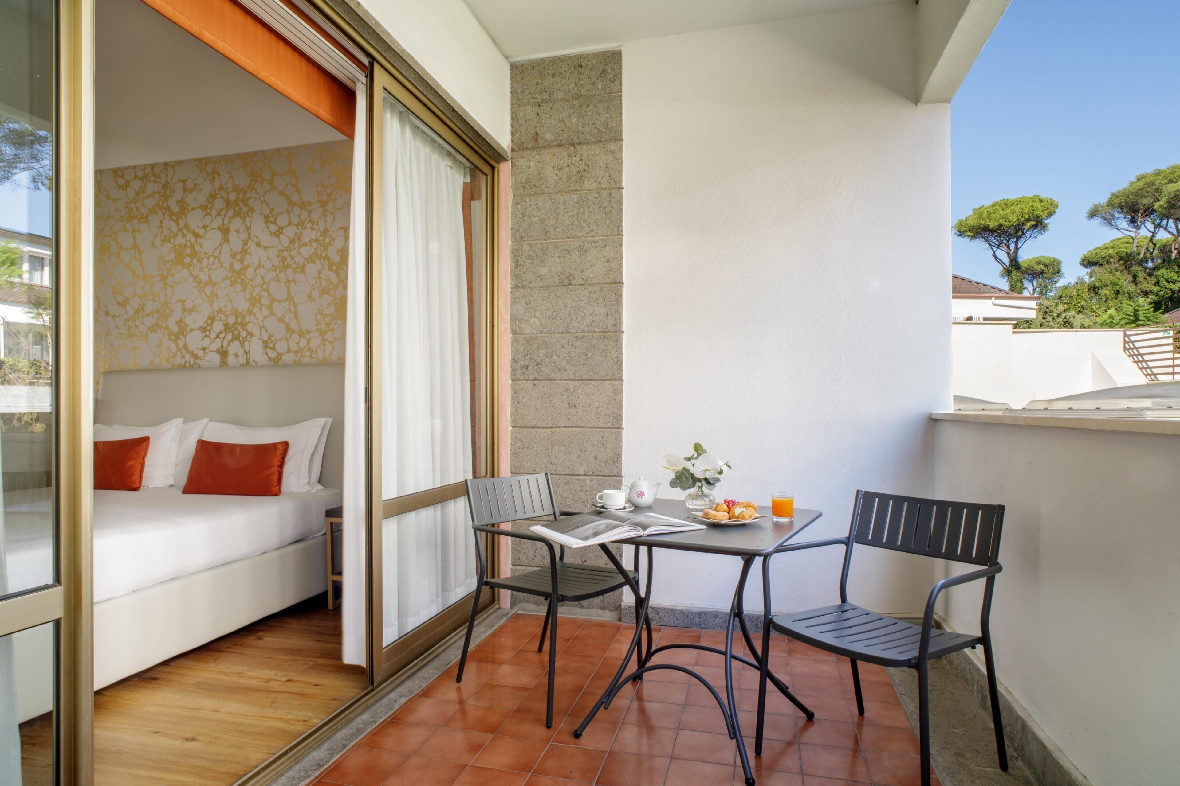 A balcony with a bed, two chairs, a table, and a small table with a cup, saucer, and flowers.