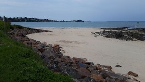 Une plage bordée d’une côte rocheuse, avec une partie sablonneuse, et d’où l’on peut apercevoir une ville lointaine de l’autre côté de l’eau.